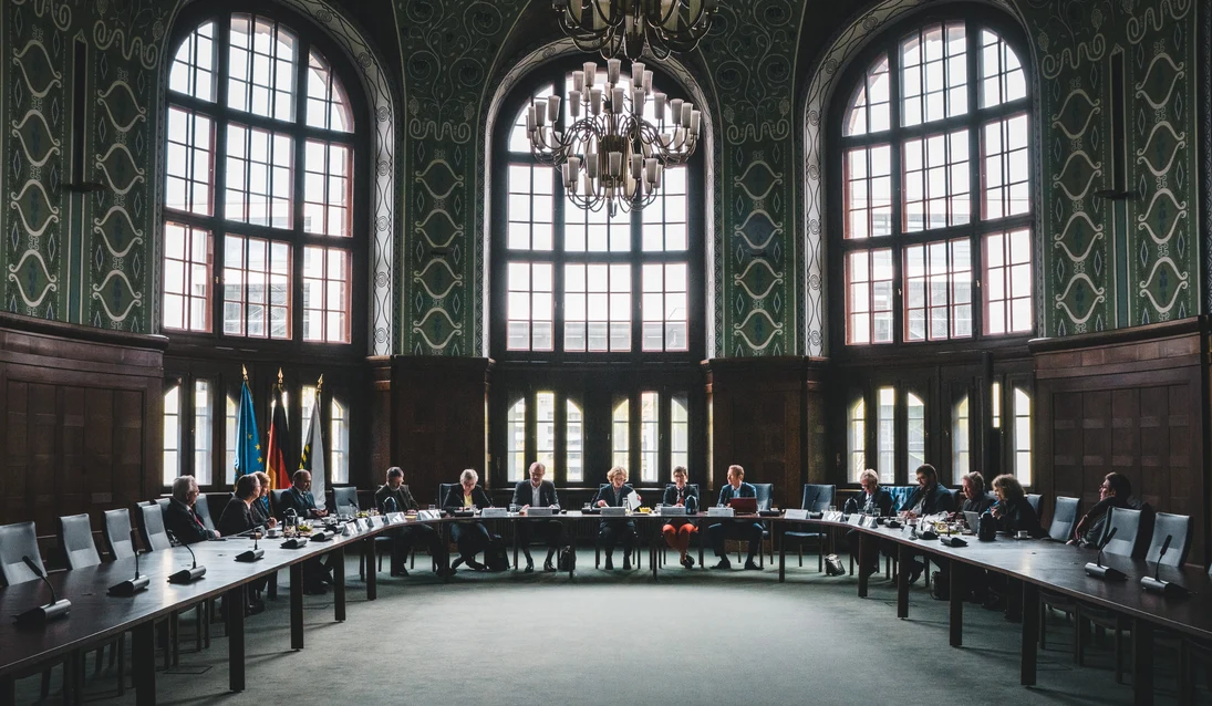 Blick in einen prunkvollen Saal, in einem langen Tisch sitzen Personen