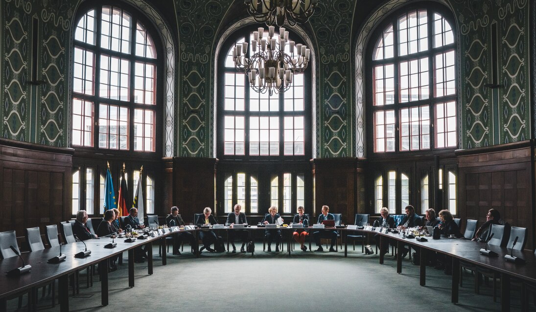 Blick in einen prunkvollen Saal, in einem langen Tisch sitzen Personen