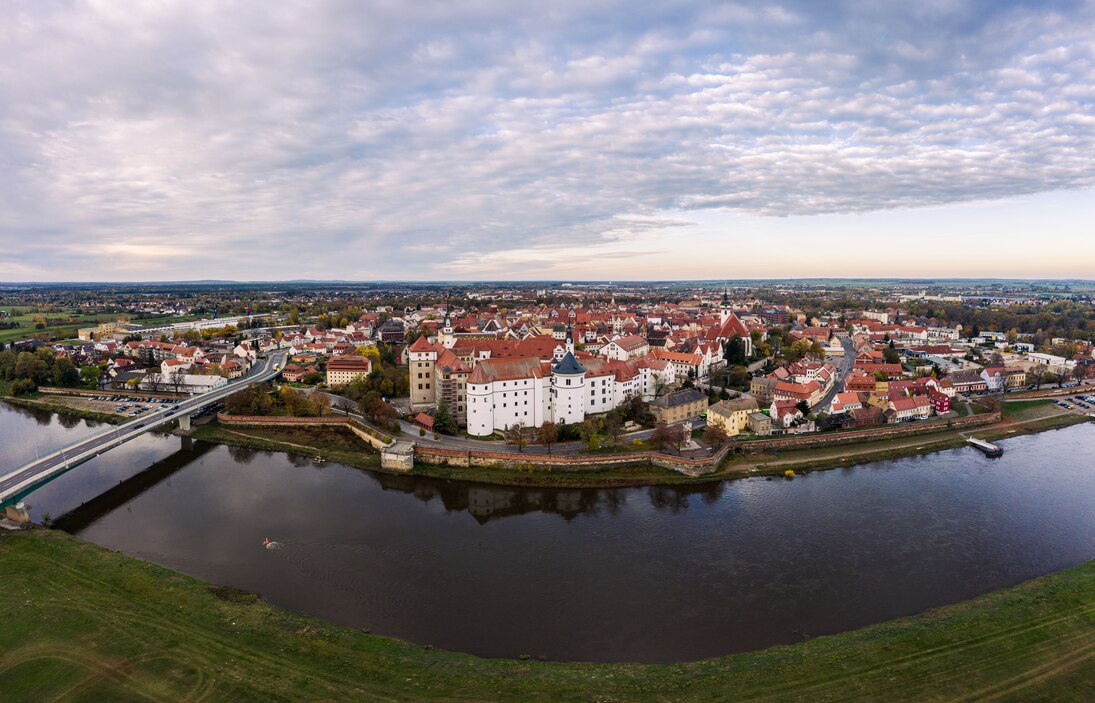 Luftbild von Torgau in Sachsen. Vorne ist ein Fluss zu sehen, dann ein Schloss und dahinter die schöne Stadt